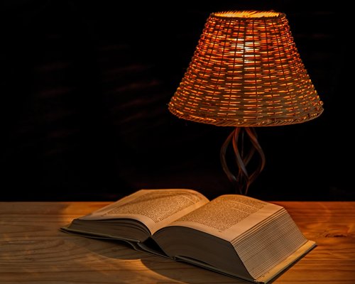 An overhead view of an open book illuminated gently by a warm yellow reading light on a nightstand